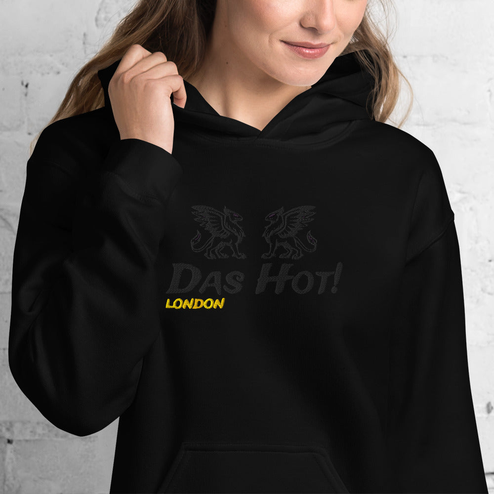 Close up view of a female model wearing the premium black unisex streetwear hoodie by Das Hot! London. The image showcases the high-density black-on-black raised embroidery of the twin heraldic griffins and the Das Hot! brand name positioned on the chest, with the contrasting yellow-gold embroidered London accent placed directly underneath the brand name.