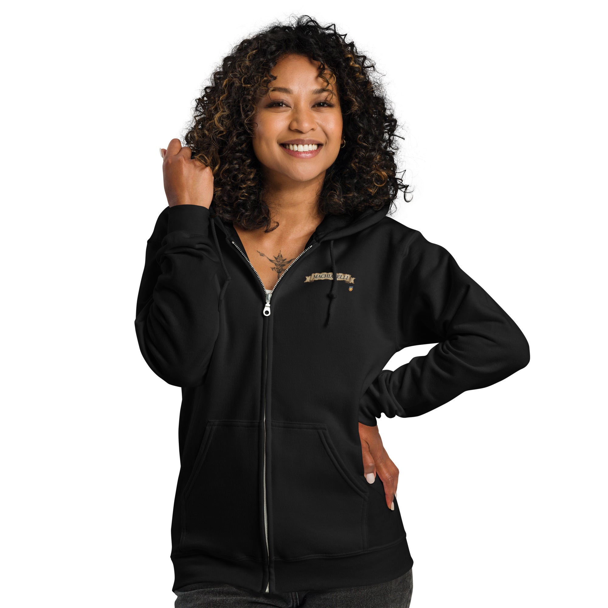 Close up portrait of a smiling female model with curly dark hair wearing the premium black zip-up streetwear hoodie by Das Hot!. The hoodie is partially unzipped, showing a silver metal zipper and the embroidered parchment scroll banner featuring the name MACHIAVELLI on the left chest against a white studio background.