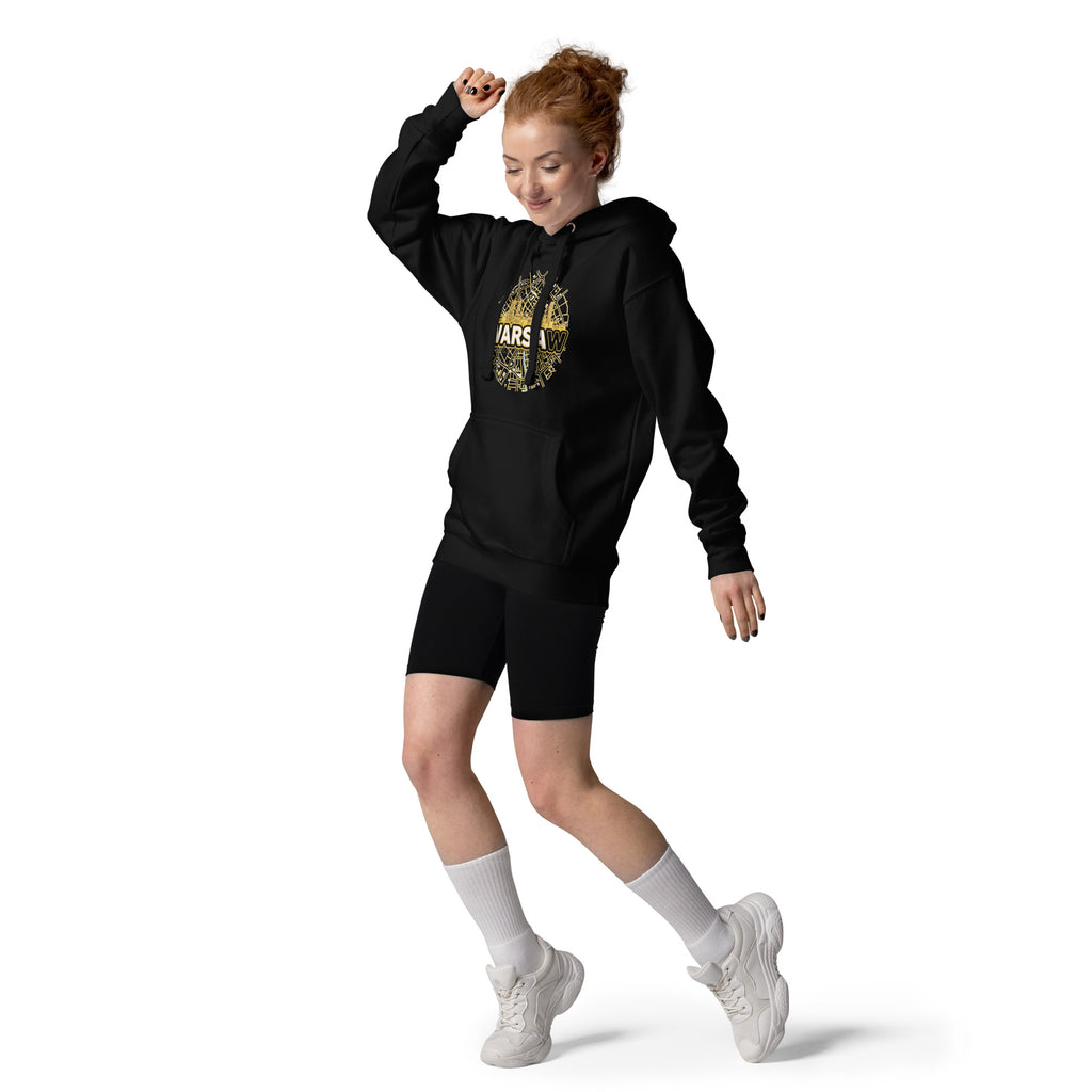 Full length view of a smiling female model with red hair captured mid-motion, showcasing the black premium unisex streetwear hoodie by Das Hot!. The hoodie features the circular gold Warsaw city map and skyscraper skyline graphic on the chest. The casual urban look is styled with black biker shorts, high white socks, and chunky white sneakers against a plain white studio background.
