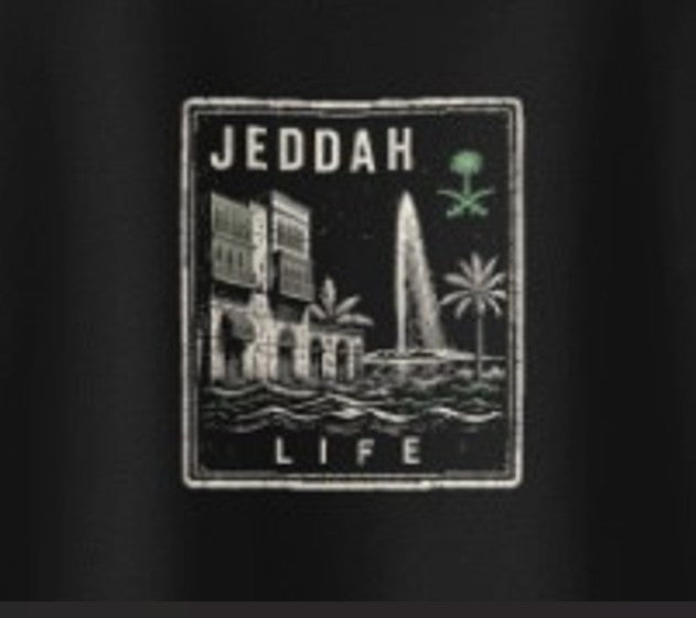 Extreme close-up of the Jeddah Das Hot! Passport™ Jeddah Life badge on the back of a black Das Hot! oversized streetwear T-shirt featuring Al-Balad architecture, the Jeddah Fountain, palm trees, and Saudi-inspired artwork.