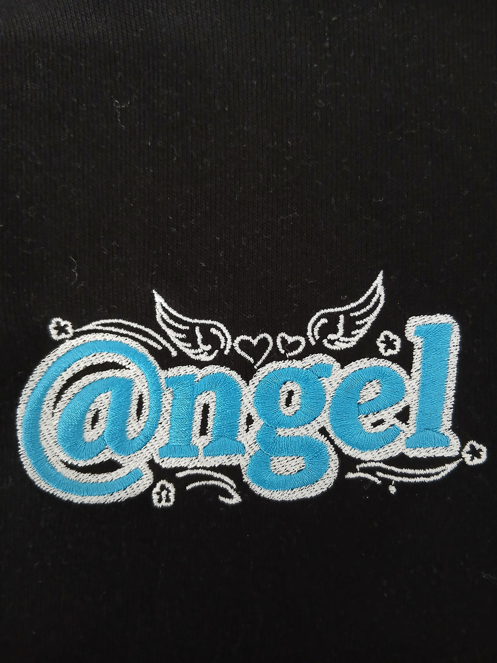 A macro close-up view of the premium graphic embroidery on the black Dashot cropped hoodie. The image showcases the heavy, high-density stitching of the stylized "@ngel" (Angel) logo in a vibrant sweet turquoise blue fill, tightly bordered by sharp white thread outlines. Intricate embroidery details are fully visible, including the stylized "at" sign, curved accent lines, small stars, and delicate winged hearts centered above the typography, all contrasting against the deep midnight black cotton-blend fabri