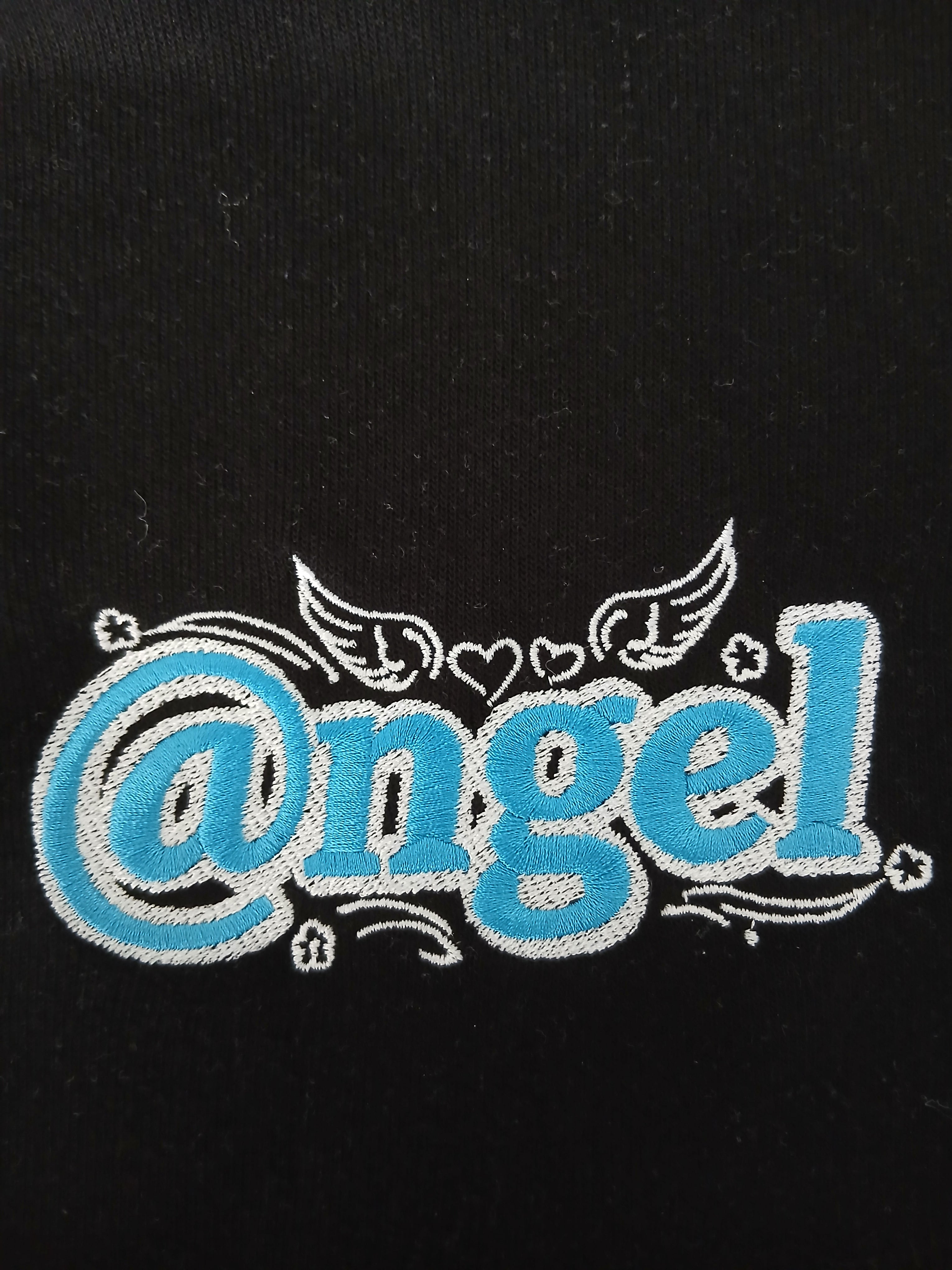 A macro close-up view of the premium graphic embroidery on the black Dashot cropped hoodie. The image showcases the heavy, high-density stitching of the stylized "@ngel" (Angel) logo in a vibrant sweet turquoise blue fill, tightly bordered by sharp white thread outlines. Intricate embroidery details are fully visible, including the stylized "at" sign, curved accent lines, small stars, and delicate winged hearts centered above the typography, all contrasting against the deep midnight black cotton-blend fabri