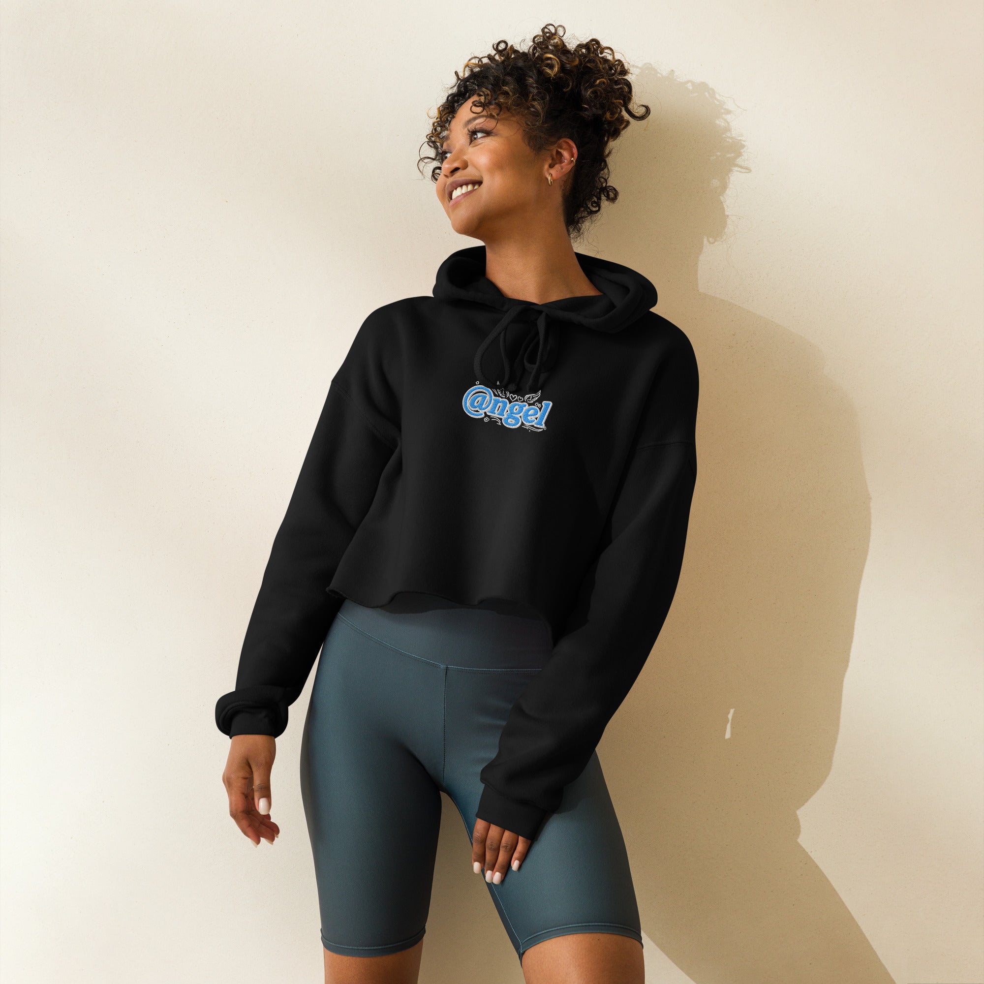 A lifestyle front view of a smiling female model wearing the black Dashot cropped hoodie. The shot clearly showcases the fit and drape of the premium fleece, featuring the high-contrast light blue and white "@ngel" (Angel) typography embroidery across the chest. The visual highlights the stylish raw-edge cropped hemline, flat black drawstrings, and relaxed drop-shoulder street