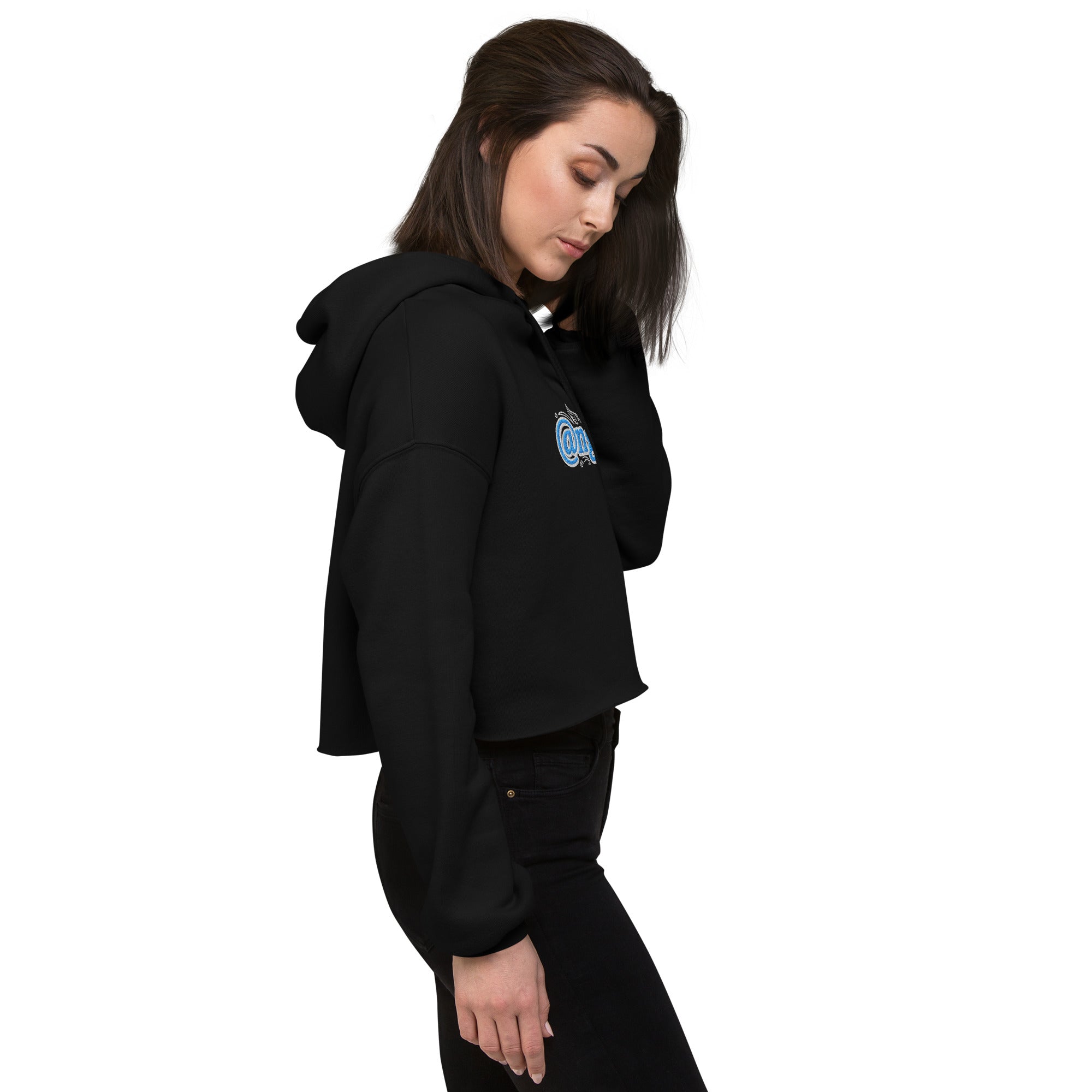 A side lifestyle view of a female model looking down while wearing the black Dashot cropped hoodie. The shot emphasizes the structured profile of the relaxed streetwear hood, drop shoulders, and thick premium fleece texture, while catching a partial side glimpse of the vibrant light blue and white "@ngel" (Angel) embroidery on the chest.