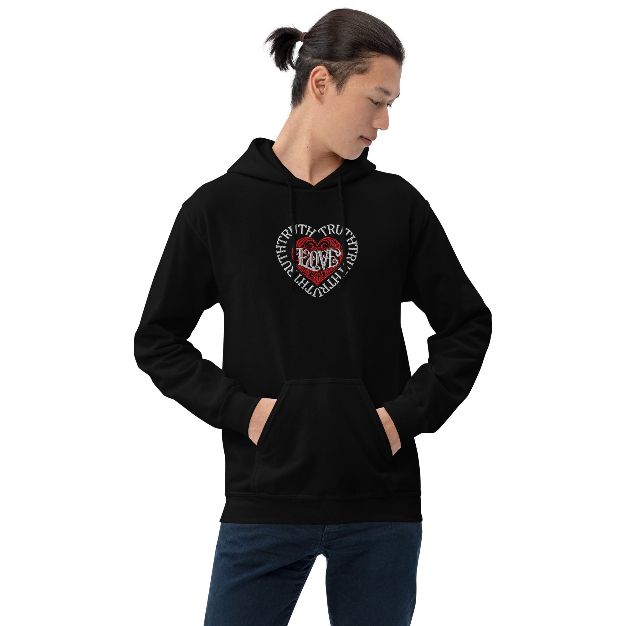 A man with a ponytail looks down in a front lifestyle view wearing a solid black Dashot hoodie, displaying a premium embroidered chest piece where white stitched text of TRUTH forms a protective heart border enclosing a red stitched tribal heart with the word LOVE at its center.
