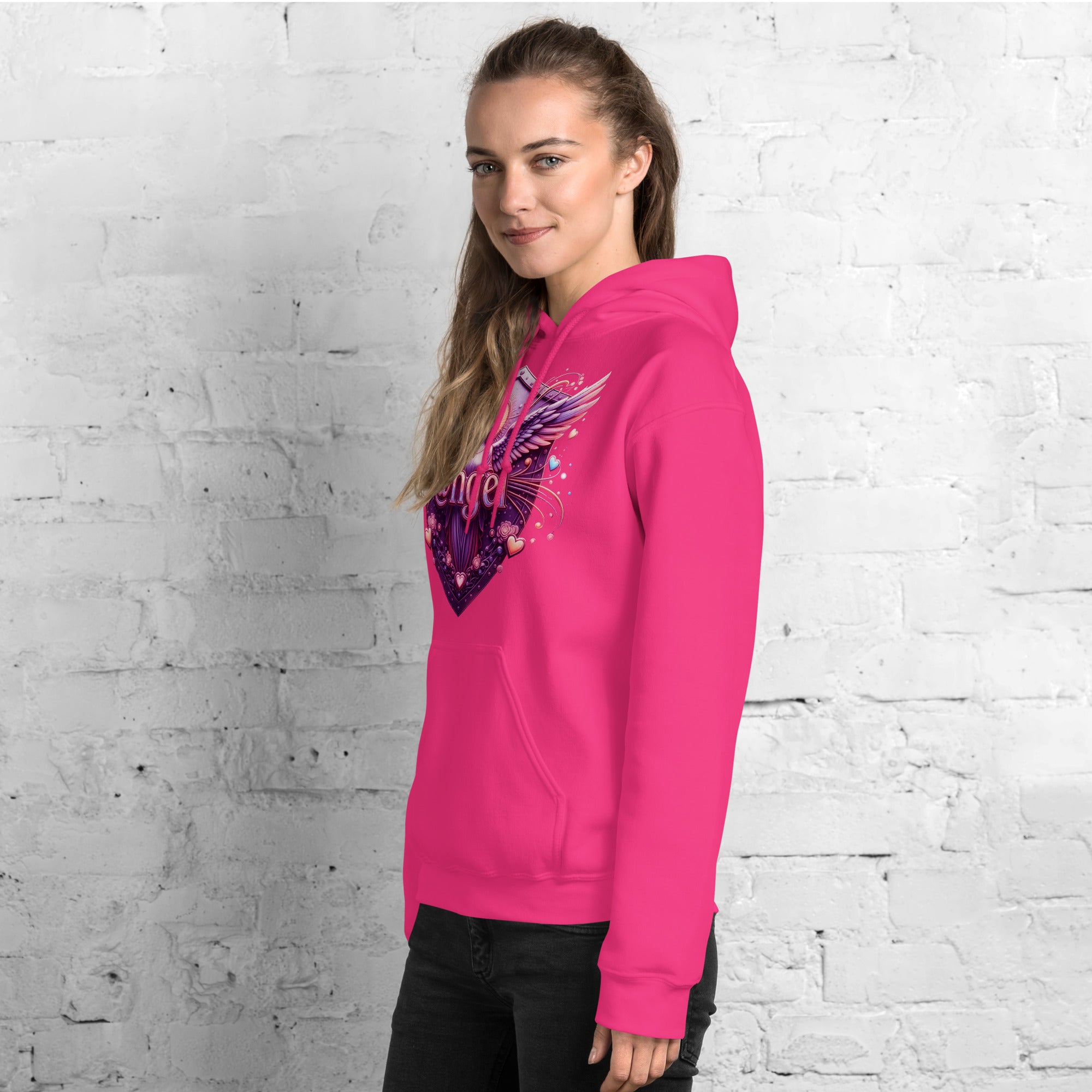 woman wearing a bright pink hoodie with an angel graphic design against a white brick wall.